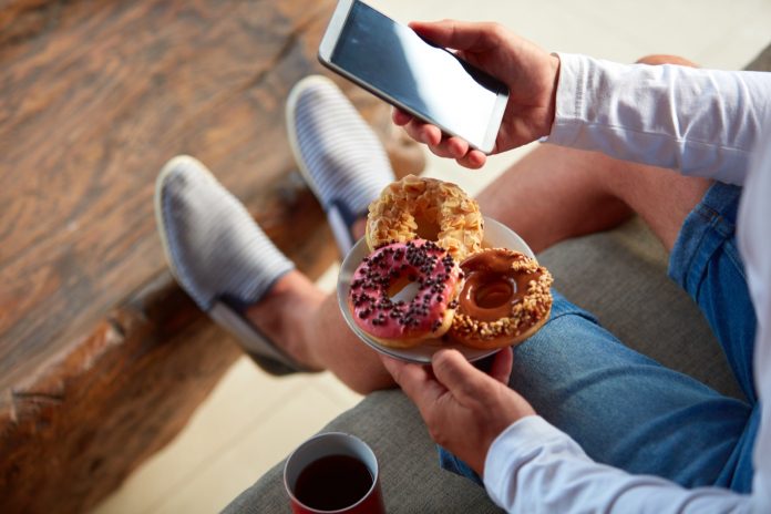 Man sitting on a terrace sofa and drinking coffee/tea, eating donuts while using cellphone.