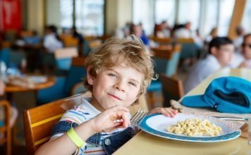 Cute healthy preschool kid boy eats pasta noodles sitting in school or nursery cafe. Happy child eating healthy organic and vegan food in restaurant. Childhood, health concept.