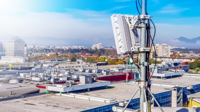 5G mobile telecommunication cellular radio network antenna on a mast on the roof broadcasting signal waves over the city on a clear sunny day with blue sky and clouds