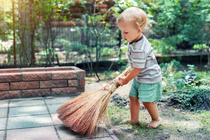 Cute adorable caucasian toddler boy playing holding broom at backyard in garden outdoors. Child little helper in t-short and shorts having fun sweeping and cleaning yard near house at countryside