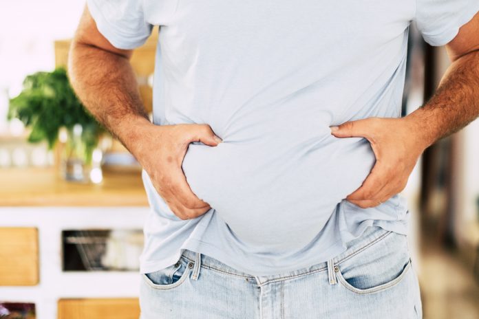 Caucasian fat man hold his belly with hands - diet and healthy lifestyle concept - junk food results - home in background and quarantine coronavirus result