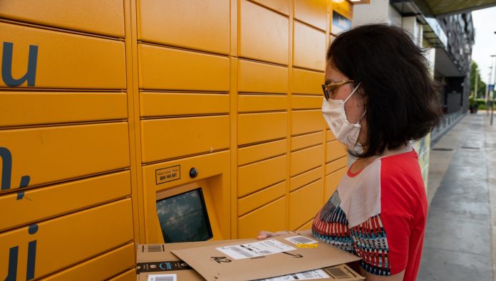 A Caucasian woman is picking up her shipment at an amazon locker. He holds the packaging with the logo in his hand and reads the communications on the screen.