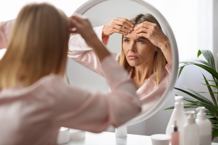 Stressed Mature Lady With Pimple On Forehead Looking In Mirror At Home