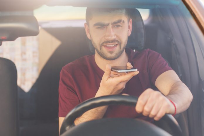 Bearded young male with thick stubble makes voice call via cell phone, drives car on high speed, risks as has distracted attention, sits in front of wheel. People, technology and driving concept