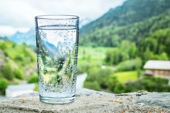 Glass of water on the stone close-up. Blurred snow mountains tops and green forests at the background.